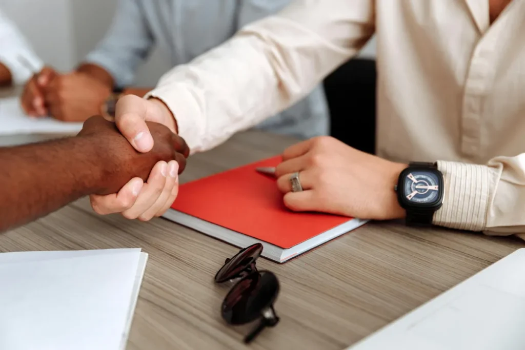 Dua eksekutif bisnis berjabat tangan di atas meja dengan dokumen perjanjian merger